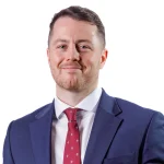 An image of Will Busby. He is smiling at the camera, wearing a blue suit with a red tie.