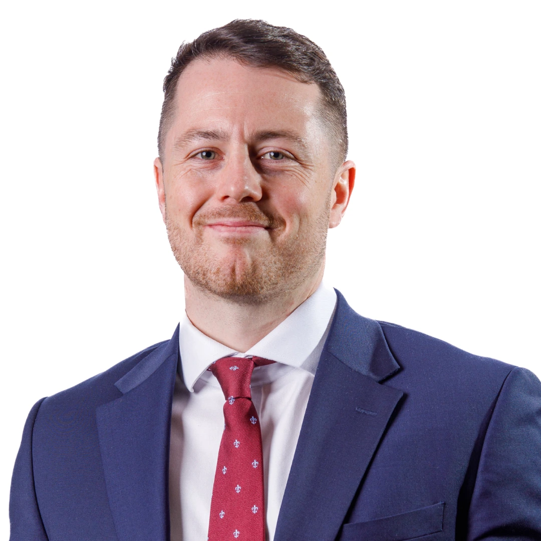 An image of Will Busby. He is smiling at the camera, wearing a blue suit with a red tie.