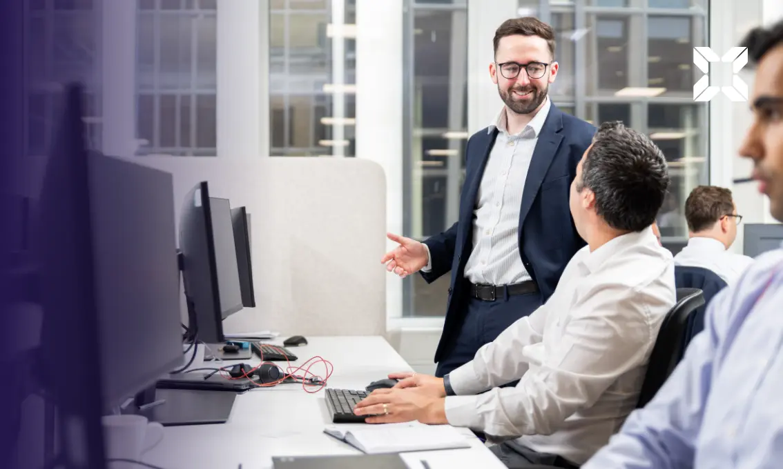 A man in a suit talking to another man sat at a desk in front of a computer.