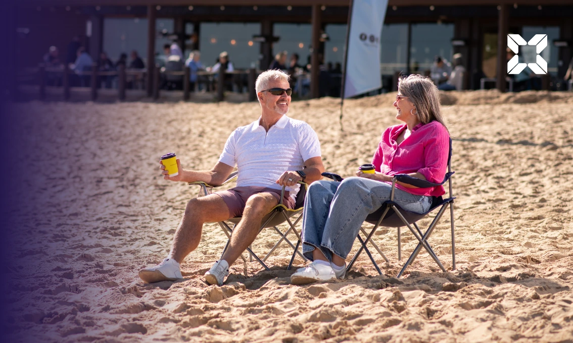 A retired couple sat on camping chairs at a sunny beach enjoying a coffee.