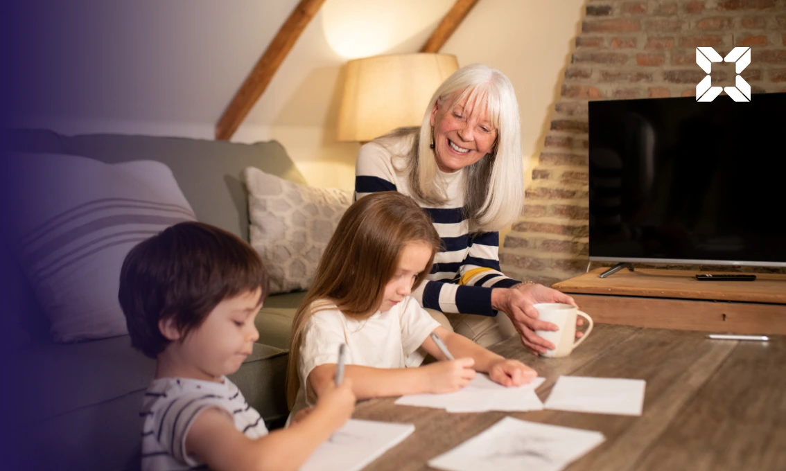 A grandmother is sat on a couch watching her two grandchildren drawing.
