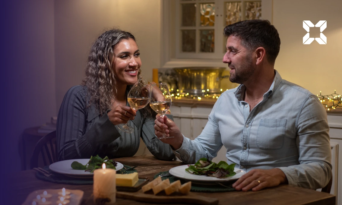 A middle aged couple are sat at a dining table enjoying a romantic meal together. They are smiling at each other and are clinking champagne glasses.