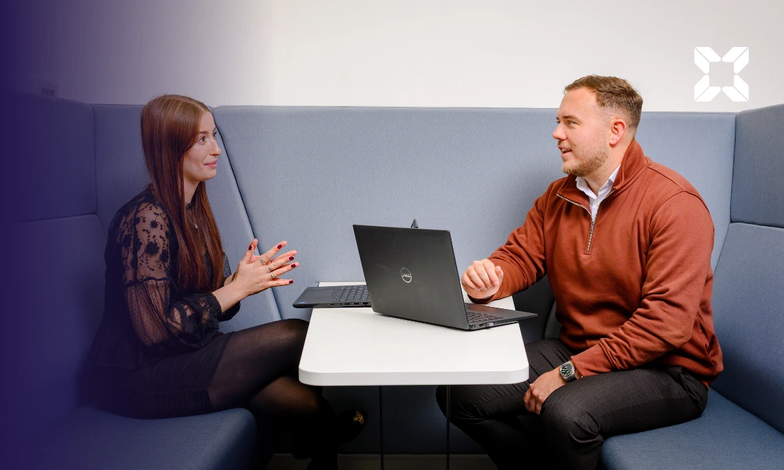 A man and a woman are sat opposite each other in a booth having a discussion. They have laptops open in front of them.