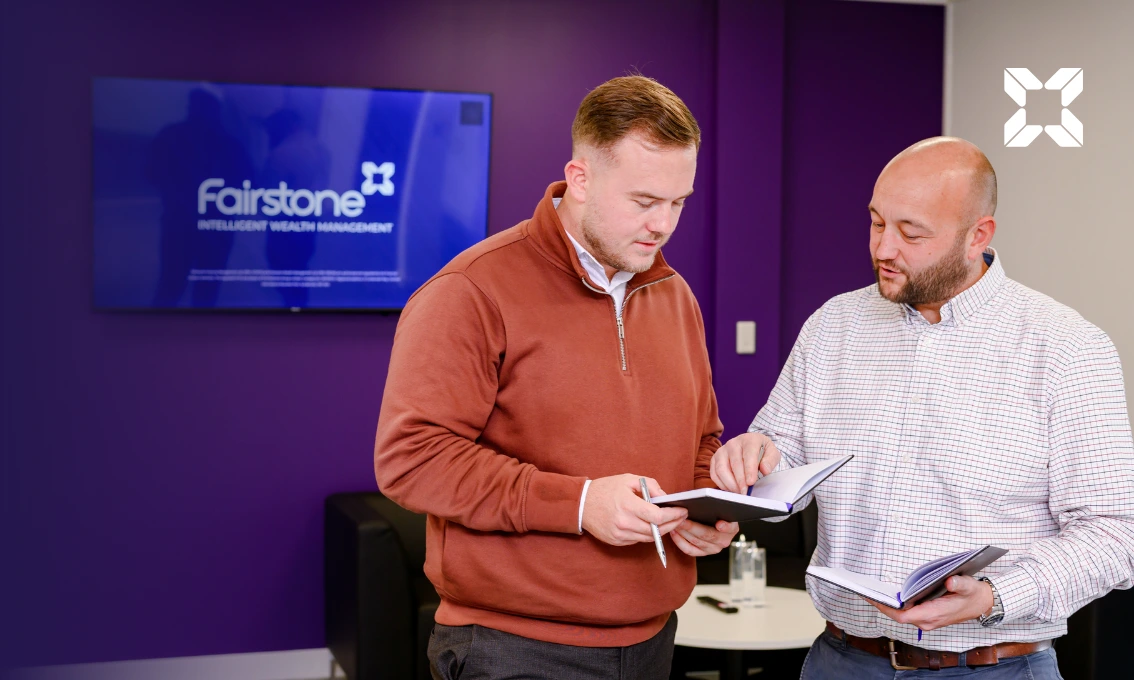 Two men are stood up in an office having a conversation and looking at their notebooks