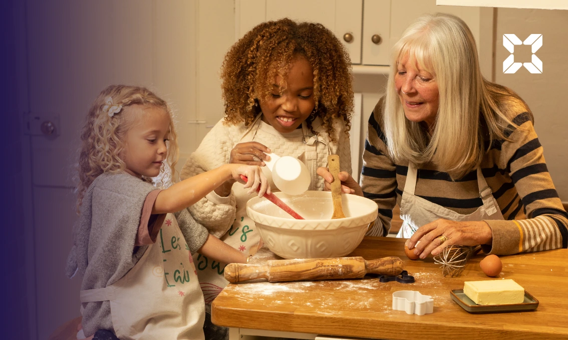 A grandmother is baking with her two granddaughters in a kitchen.