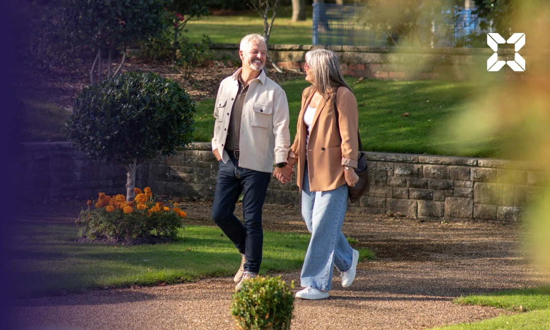 A middle aged couple are walking through a park holding hands. They are smiling at each other.