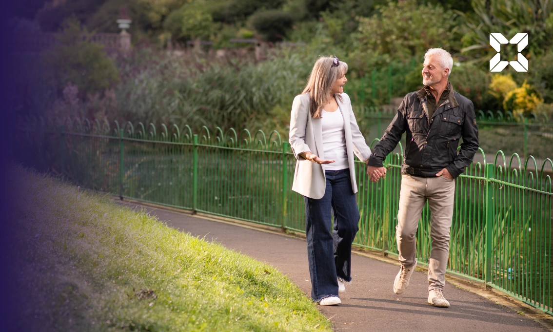 A middle aged couple are walking through a park, holding hands and talking to each other.